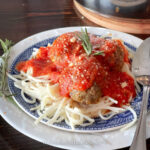 A plate of spaghetti topped with meatballs and rich Instant Pot homemade marinara sauce, garnished with a sprig of rosemary. The dish is served on a white and blue patterned plate with a metal spoon beside it.