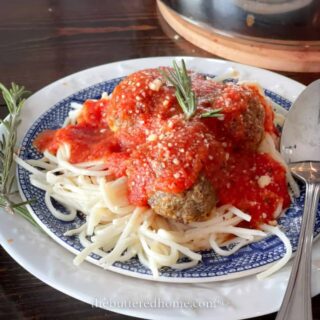A plate of spaghetti topped with meatballs and rich Instant Pot homemade marinara sauce, garnished with a sprig of rosemary. The dish is served on a white and blue patterned plate with a metal spoon beside it.