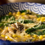 A close-up of a Butternut Squash Soup with Bacon in a blue patterned bowl, featuring spinach, corn, white beans, grated cheese, and a brown garnish. The background is softly blurred. The text "thebutteredhome.com" is visible at the bottom of the image.