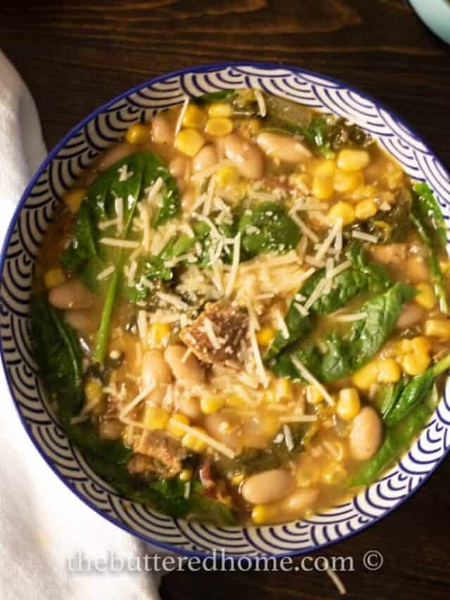 A bowl of soup containing white beans, corn, spinach, and grated cheese, served in a blue and white patterned bowl. The soup is garnished with shredded cheese on top.