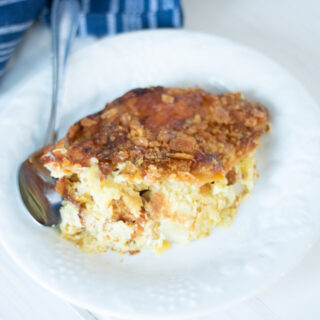 A slice of creamy Southern Yellow Squash Casserole topped with a golden, crispy crust rests on a white plate. A metal spoon sits beside it, ready to serve. A folded blue-striped cloth is partially visible in the background.