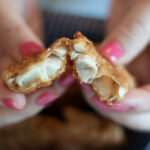 Hands with pink nail polish hold a piece of Southern Fried Fish, revealing its flaky, white interior. The background is slightly blurred.