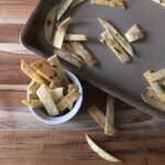 Homemade tortilla strips are scattered on a baking tray and a wooden surface. Some strips are placed inside a small white bowl next to the tray.