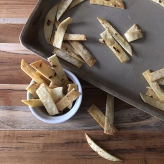 Homemade tortilla strips are scattered on a baking tray and a wooden surface. Some strips are placed inside a small white bowl next to the tray.