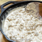 A cast iron skillet filled with homemade Southern sausage gravy is being stirred with a wooden spatula. A biscuit sits on a wooden board in the background, adding to the rustic charm. The image is elegantly watermarked with "thebutteredhome.com".
