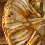A close-up of a homemade pear tarte showcases its rustic charm with a golden, flaky crust. The pear slices are neatly arranged in a circular pattern, appearing glazed for a shiny look. Set on a wooden surface, the delicacy is highlighted by the subtle touch of a text watermark.