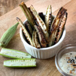 A small white ramekin is filled with oven-roasted okra fries on a wooden surface. Beside it, fresh okra pods accompany a bowl of creamy dip topped with seasoning. The website "thebutteredhome.com" is visible at the bottom.