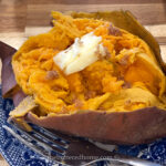A baked sweet potato, perfectly cooked using an Instant Pot, is split open with a pat of melted butter and a sprinkle of brown sugar on top. It rests invitingly on a blue and white patterned plate, with a fork beside it for the first delicious bite.