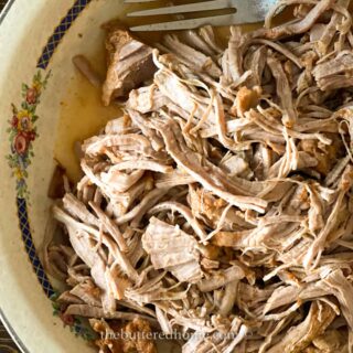 A close-up of moist, tender Instant Pot BBQ Pulled Pork Tenderloin in a floral-patterned bowl, with a metal fork partially visible at the top and savory juices pooling at the bottom.