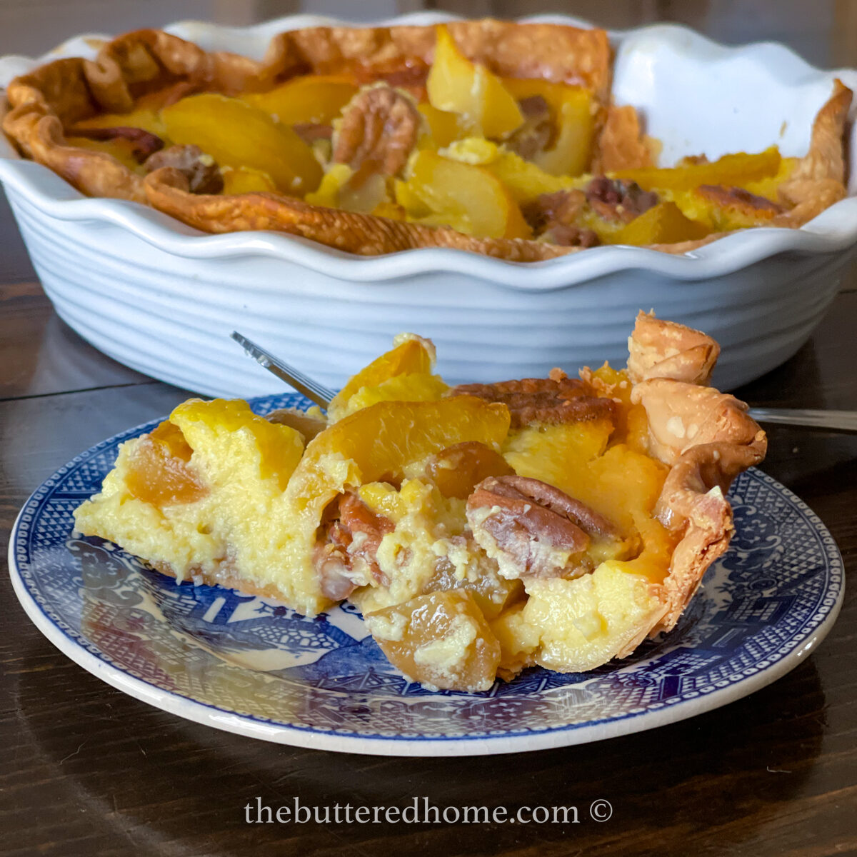 A slice of peach and pecan custard pie on a blue and white plate is in the foreground, with the full pie in a white fluted dish in the background. The pie boasts a golden crust topped with juicy peaches and crunchy pecans, accompanied by a fork on the plate.