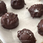 Several round chocolate truffles, some topped with coarse salt, rest on white parchment paper. One is cut open to reveal its filling—a decadent treat among the 60 Traditional Christmas Cookie Recipes to Sweeten your Holiday. "thebutteredhome.com" appears below.