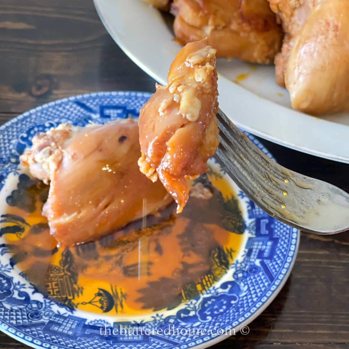 A fork holds a piece of Asian BBQ Chicken Thighs above a blue and white plate with orange sauce. More chicken pieces are on a larger white plate in the background, set on a dark wooden table.