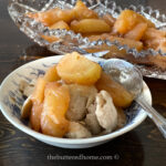 A bowl filled with apple dumplings topped with thick apple slices, delicately seasoned with cinnamon, sits invitingly beside a larger dish containing more of the apple mixture. Prepared like baked apples in the Instant Pot, a spoon rests ready in the bowl on the wooden table's edge.