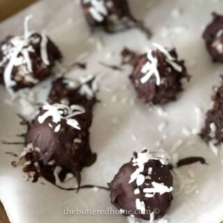 Chocolate-covered treats topped with shredded coconut are arranged on parchment paper, evoking the charm of traditional Christmas cookie recipes. The image is watermarked with "thebutteredhome.com.