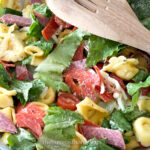 A close-up of an Italian Tortellini Salad showcases tortellini, pepperoni, salami, lettuce, cherry tomatoes, and shredded cheese. A wooden spoon is positioned in the top right corner. The image bears a watermark reading "thebutteredhome.com.