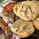 Close-up of Priesters Cinnamon Fiddlestick Cookies stacked on a cooling rack, their aromatic spices tempting your senses. A package of pecans and a nut-covered candy bar rest on a cloth with red stripes in the background, completing this cozy scene.