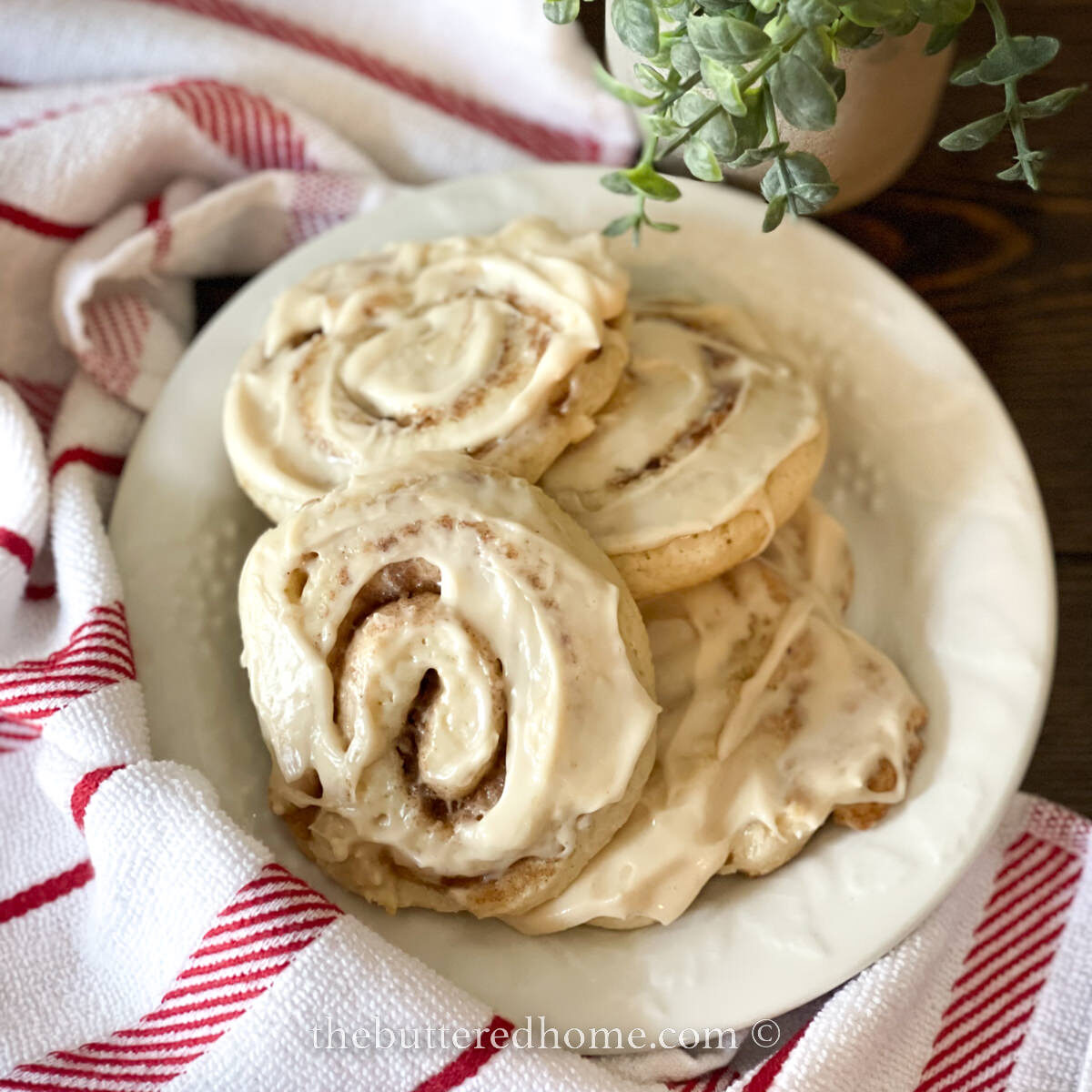 A plate of cinnamon rolls topped with icing, resting on a red and white striped cloth, could easily find its place among 25 easy and traditional Christmas recipes. A small plant in a pot is partially visible in the background.