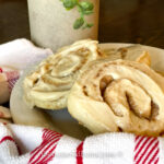 A plate with two frosted cinnamon roll cookies sits on a table, a red and white striped towel peeking beside it. In the background, a leafy plant and a sleek cylindrical container add to the cozy setting.