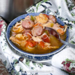 A bowl of gumbo, reminiscent of easy jambalaya, features shrimp, sausage, shredded chicken, and vegetables in a savory broth. The blue and white bowl rests on a floral-patterned fabric with a metal spoon inside.