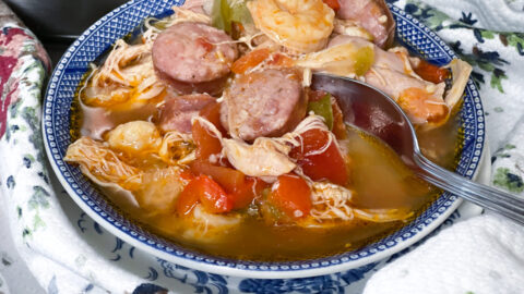 A bowl of gumbo, reminiscent of easy jambalaya, features shrimp, sausage, shredded chicken, and vegetables in a savory broth. The blue and white bowl rests on a floral-patterned fabric with a metal spoon inside.