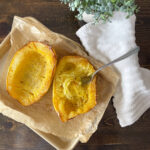 A baking tray with roasted spaghetti squash halves on parchment paper, primed for a delectable Spaghetti Squash Gratin. A fork rests in one half, pulling out delicate strands. A white cloth and a small plant elegantly adorn the dark wood table, setting a cozy culinary scene.