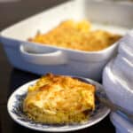 A close-up of a cheesy casserole, ranked among the top 75 Thanksgiving side dish recipes, sits on a blue and white plate with a fork. The golden-brown layers look deliciously inviting. In the background, the rest of this holiday favorite rests in a white baking dish.