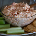 pimento cheese in a clear bowl with crackers and celery on a plate.