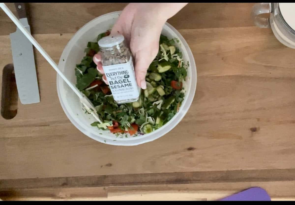 Sprinkling bagel seasoning on salad greens in a white bowl.