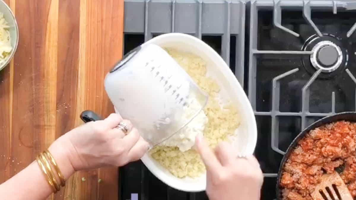 Adding sour cream mixture to the riced cauliflower in a white bowl.