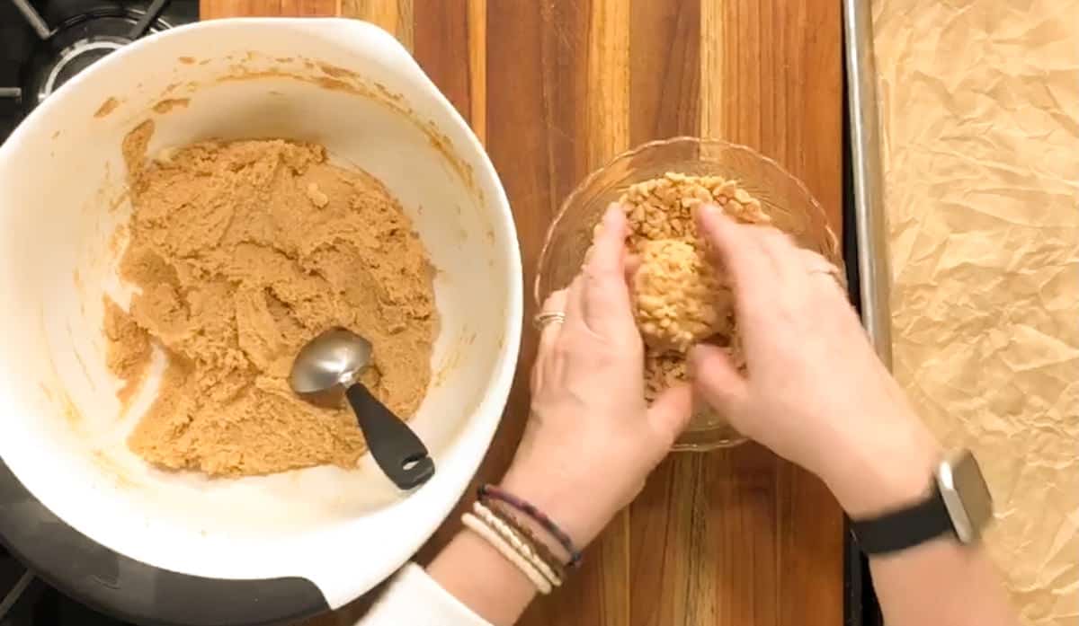A person's hands are shown forming a ball with a peanut butter mixture, beside a mixing bowl on a wooden surface, likely during a cooking or baking process.