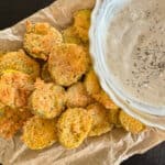 A plate of breaded and fried pickles served with a side of creamy dip garnished with herbs.