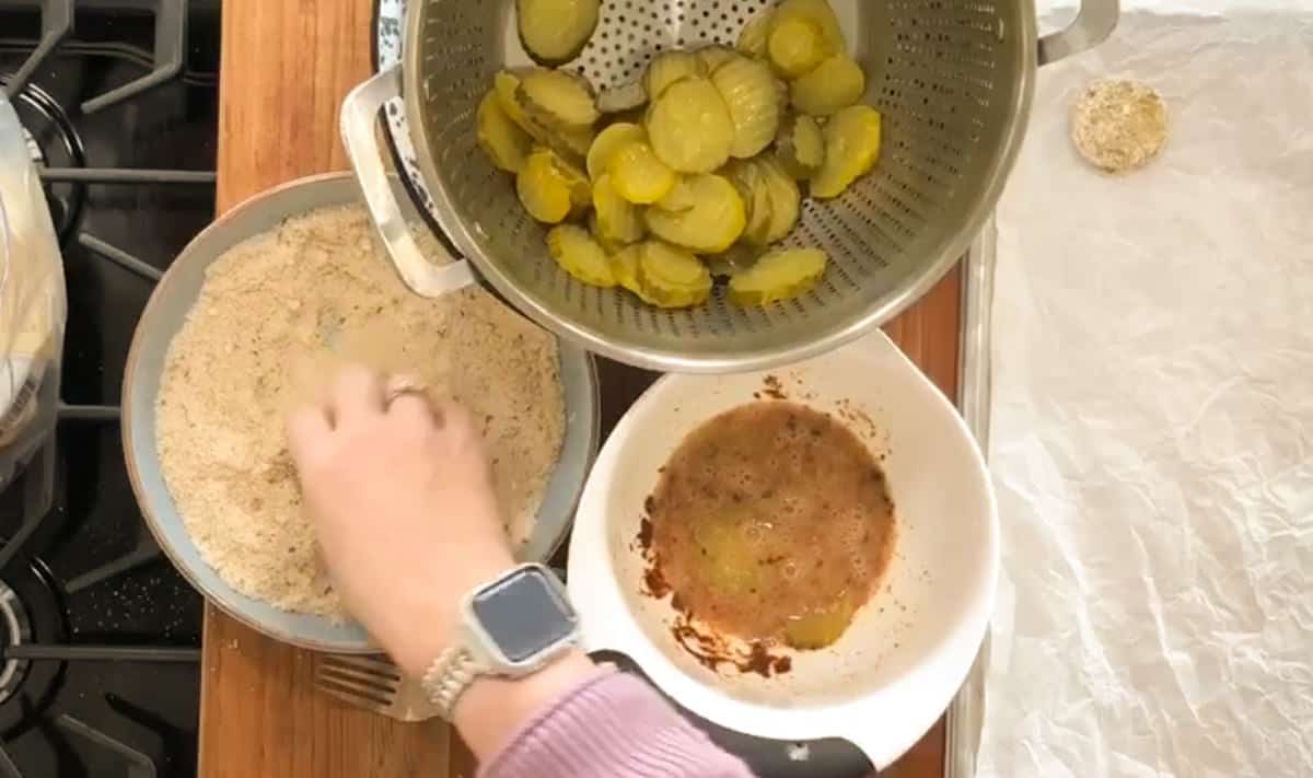 A person's hand is breading a piece of food next to a colander full of sliced pickles on a kitchen counter.