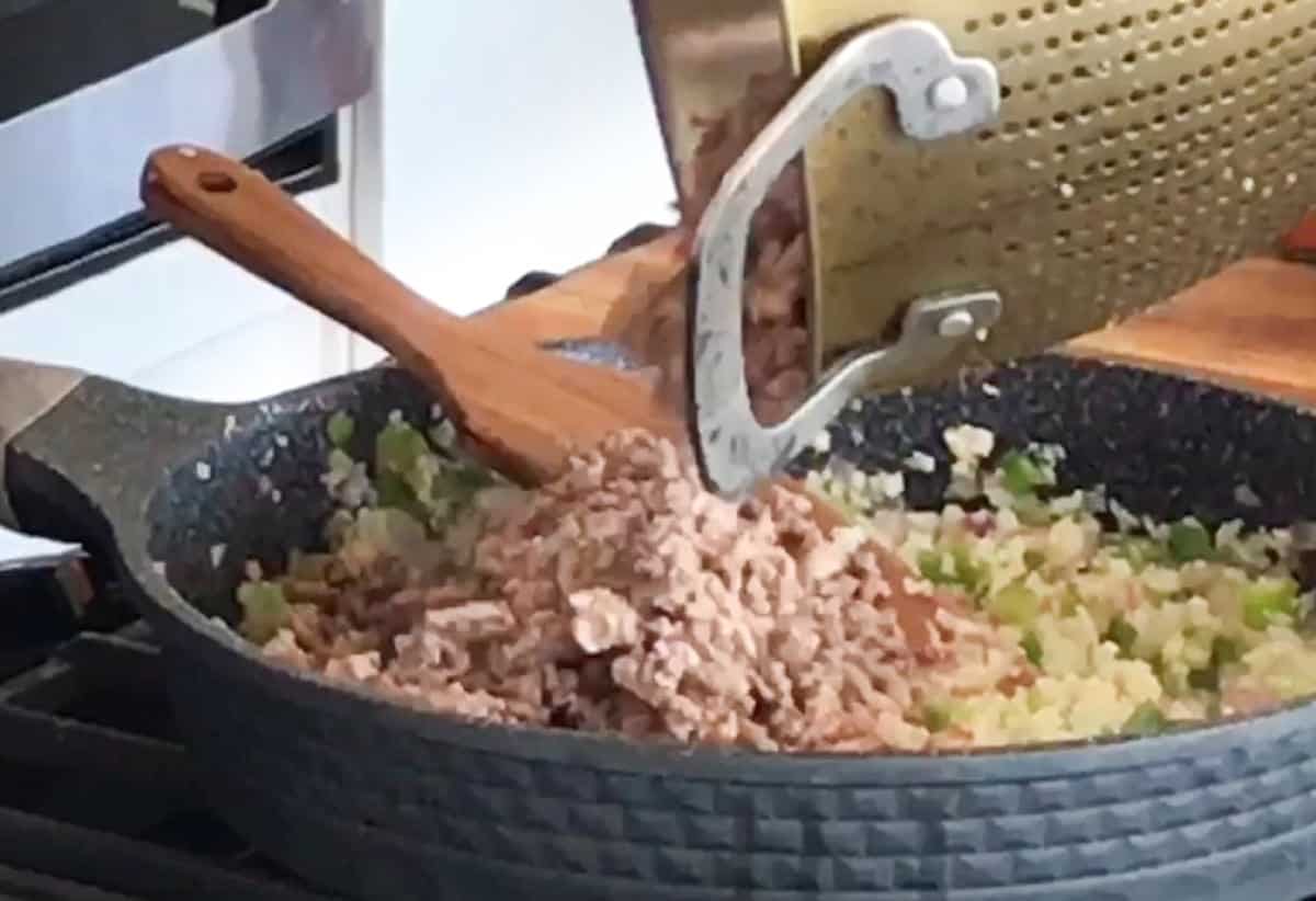 Preparing a savory dish: ground meat being added to diced vegetables in a frying pan, with a wooden spoon and a perforated kitchen tool resting on the countertop nearby.