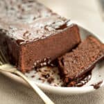 A chocolate cake with glossy icing on a plate, with one slice partially cut and a fork on the side.