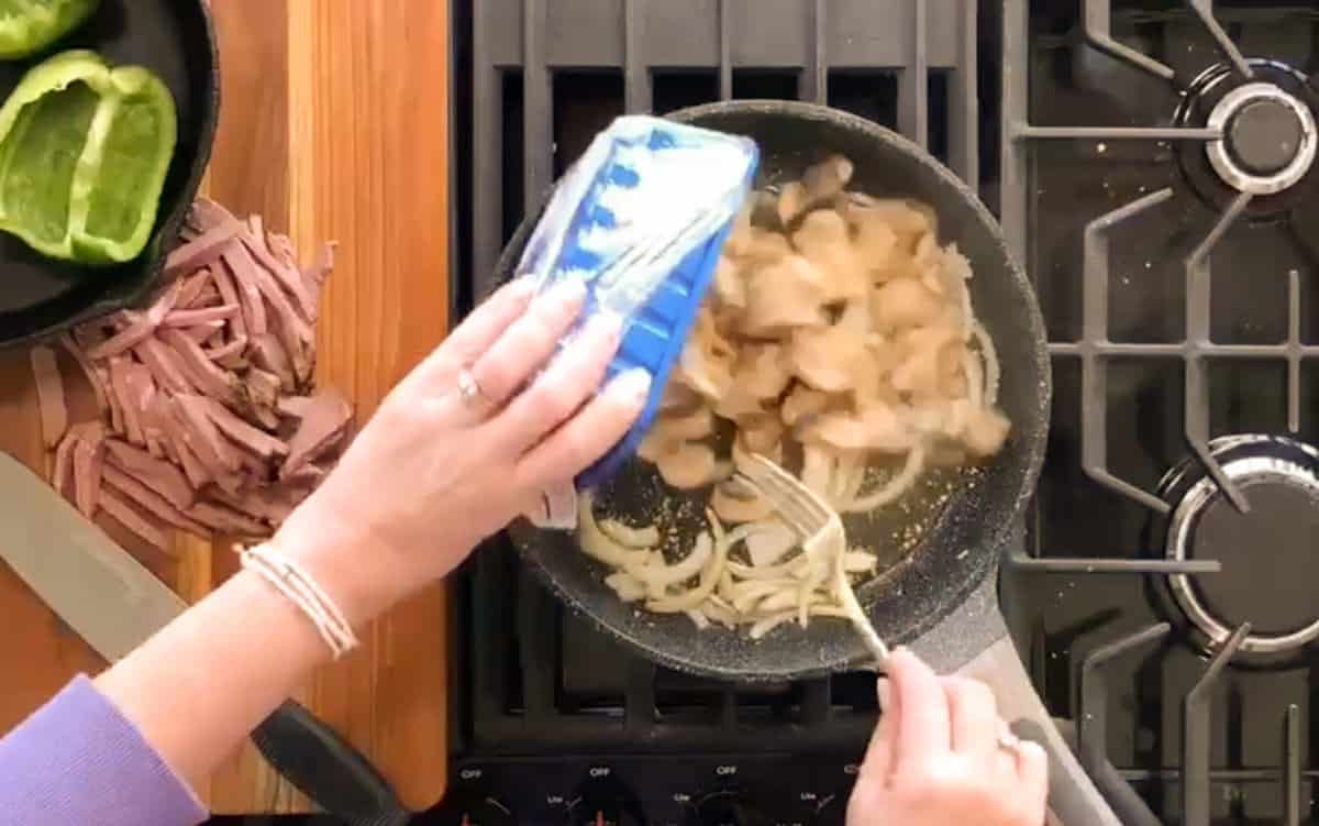 A person sautéing ingredients in a frying pan on a stove while adding seasoning from a blue container.