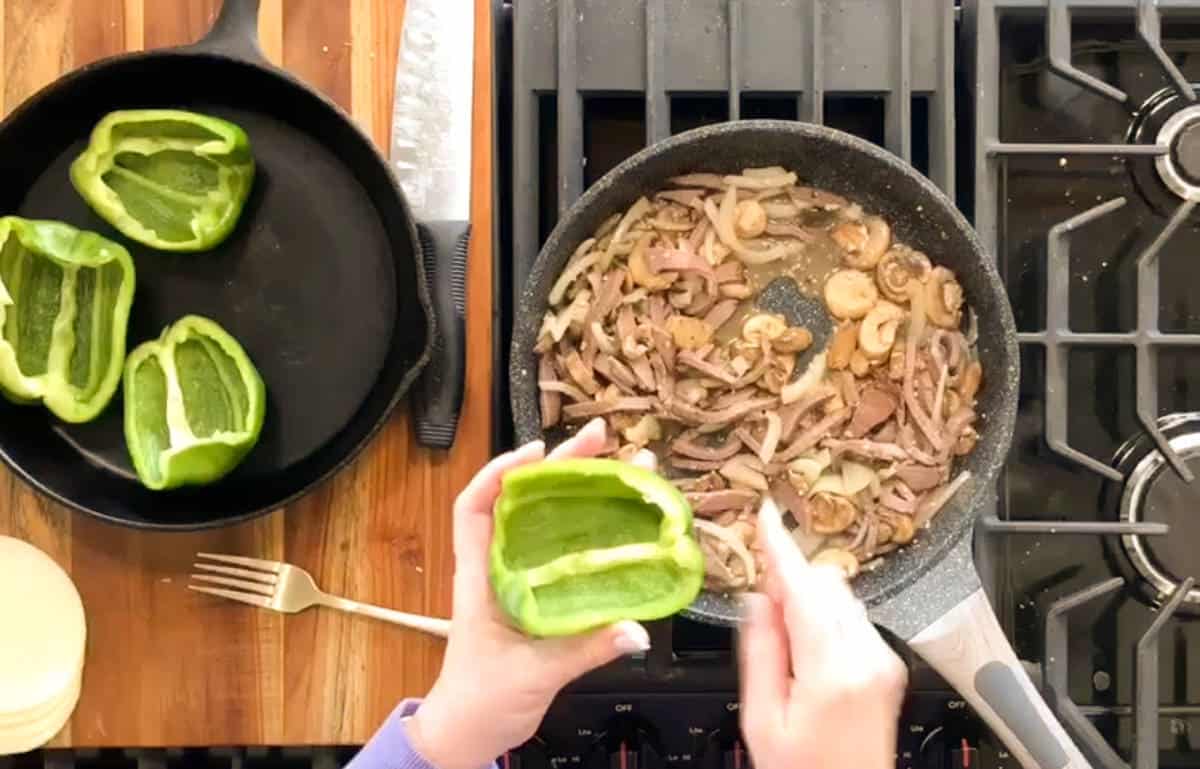 Sautéing mushrooms, sliced beef and onions in a pan on a stove with green bell peppers prepared on the side.