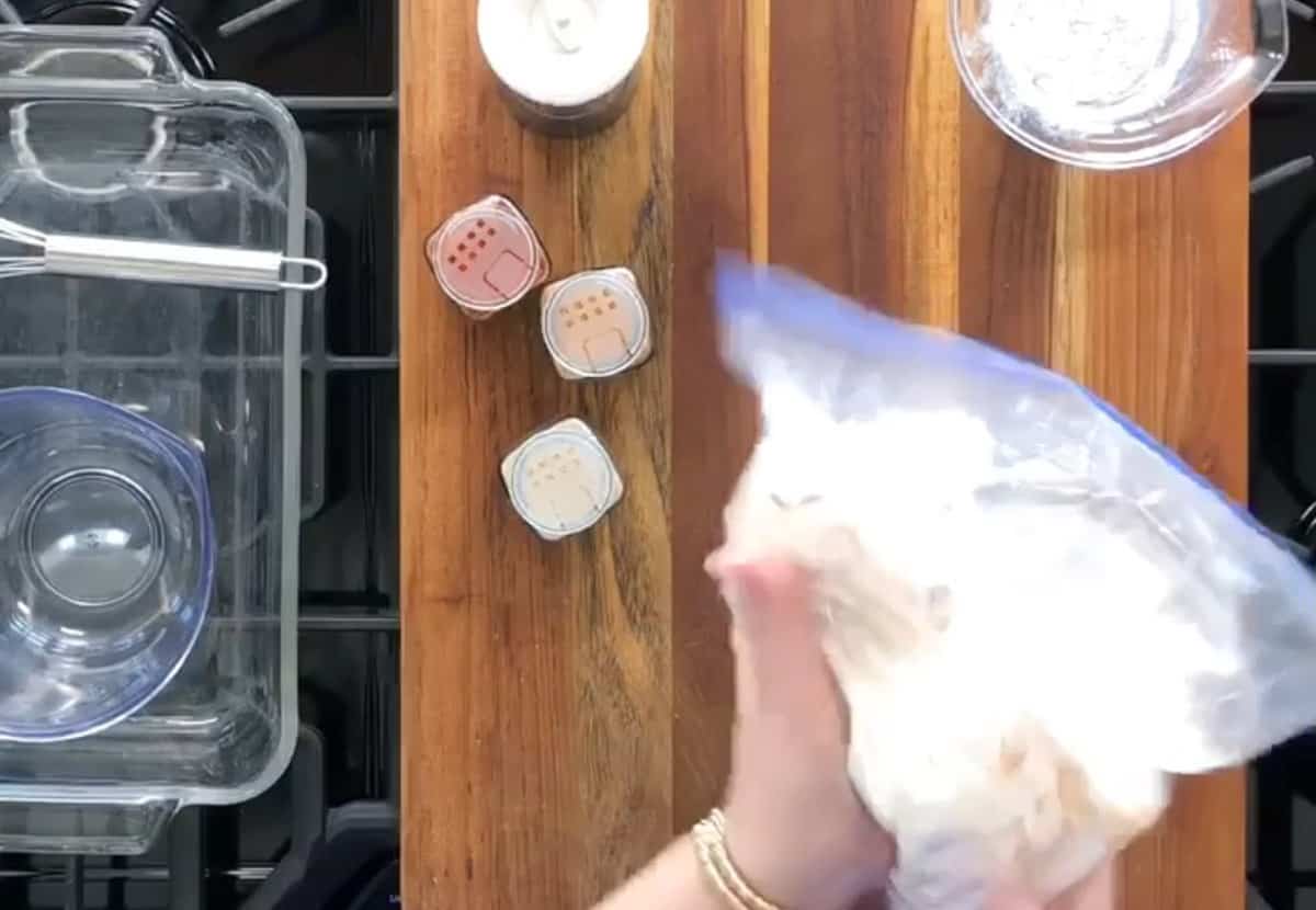 A person's hand holding a plastic bag filled with a white substance over a wooden board with containers of ingredients and kitchen utensils nearby.