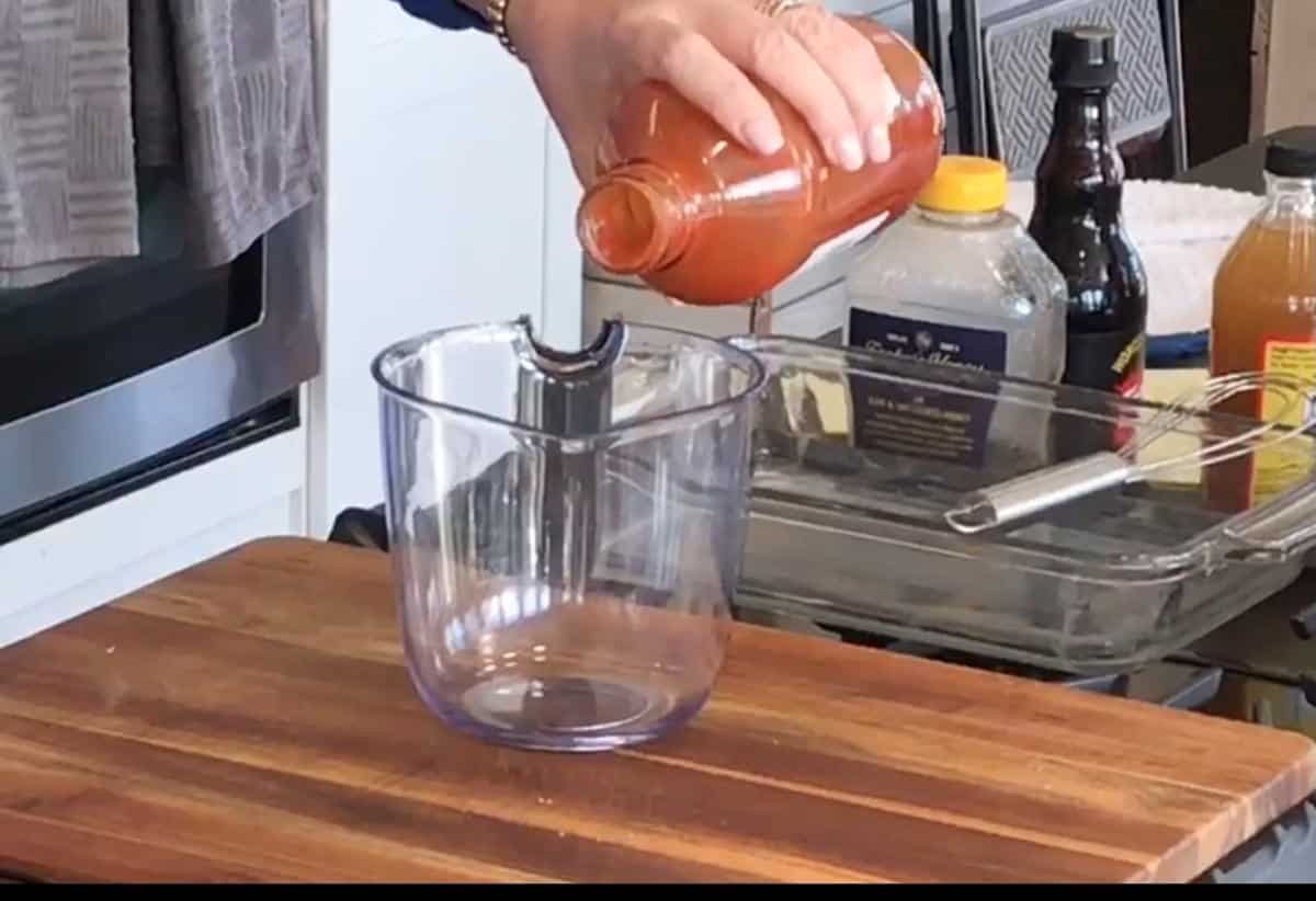A person is pouring ketchup from a bottle into a clear measuring cup on a wooden cutting board.