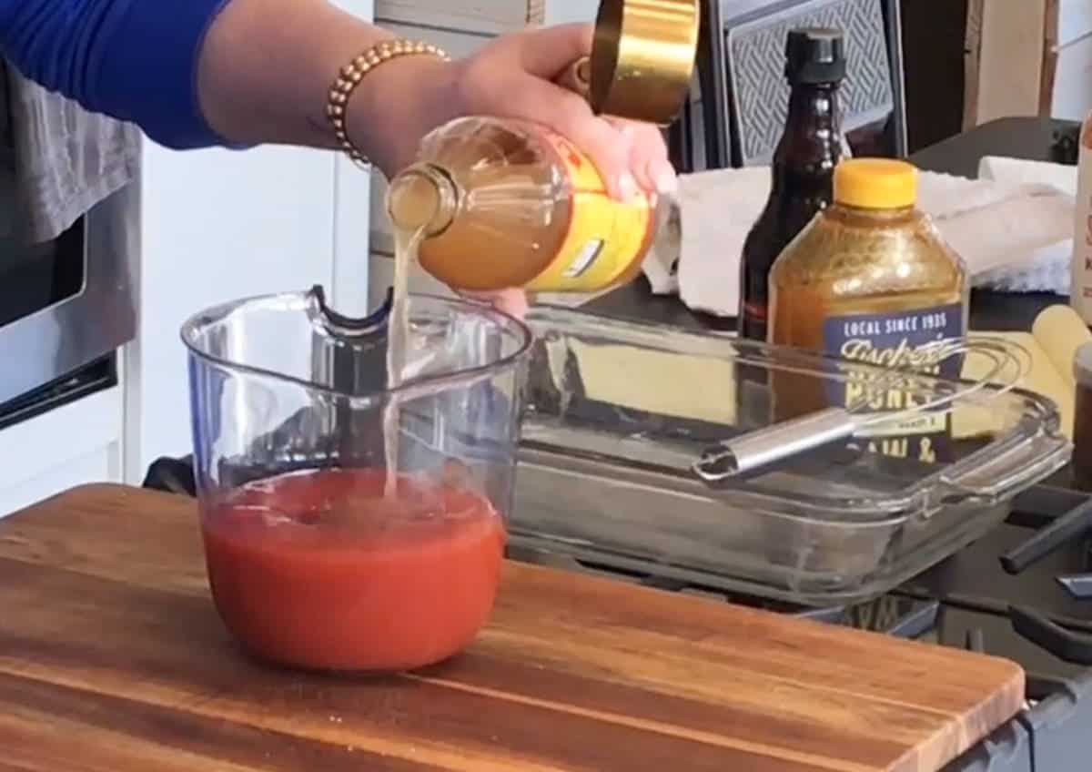 A person is pouring apple cider vinegar into a glass measuring jug with a red mixture, likely in the process of making a sauce or marinade, with various other ingredients and utensils on the countertop.
