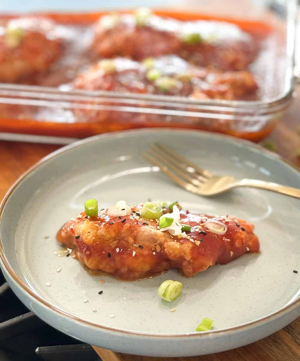 A single glazed chicken breast garnished with sliced green onions on a beige plate, with more chicken in a glass dish in the background.