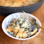 A small bowl with a blue and white pattern contains a serving of baked spinach artichoke dip. The dip is creamy, topped with melted cheese, and a fork rests inside the bowl. A black baking dish is partially visible in the background.