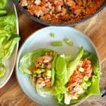 A top-down view of a meal featuring lettuce wraps filled with ground meat and diced vegetables, accompanied by a bowl of additional filling and chopped green onions on a wooden table.