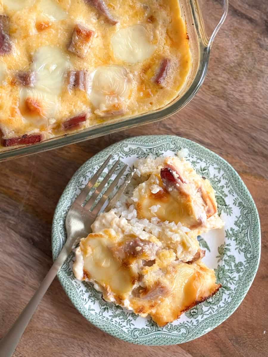 A baked dish with melted cheese and chunks of meat sits in a glass baking dish on a wooden table. A portion has been served on a decorative plate with a fork. The plate contains layers of rice, cheese, and meat.