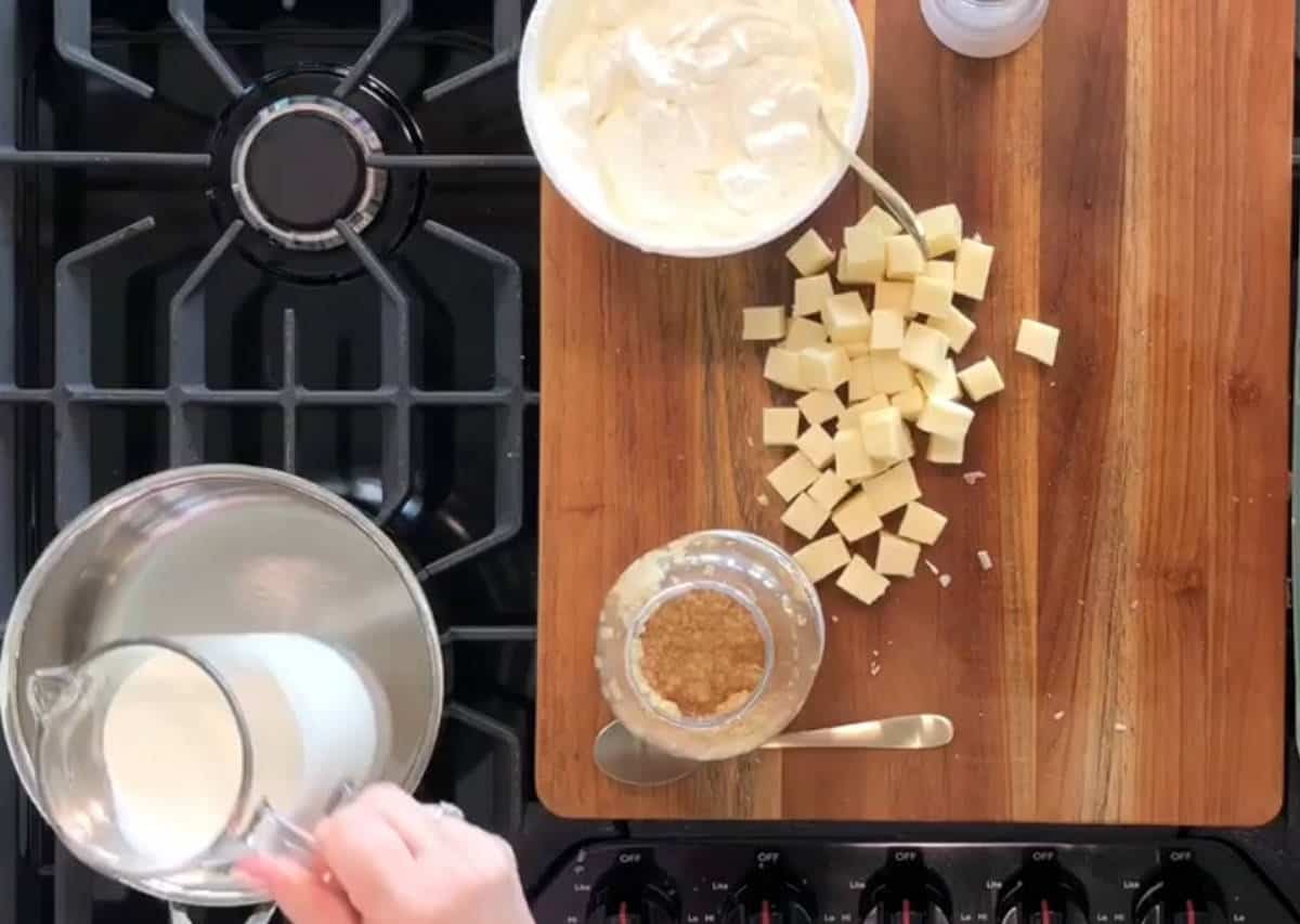 A person's hand pouring milk from a glass jug into a stainless steel pot on a stovetop. On the wooden cutting board next to the stovetop, there are cubes of cheese, a bowl with a spoon and white mixture, and a jar containing a brown substance with a spoon.