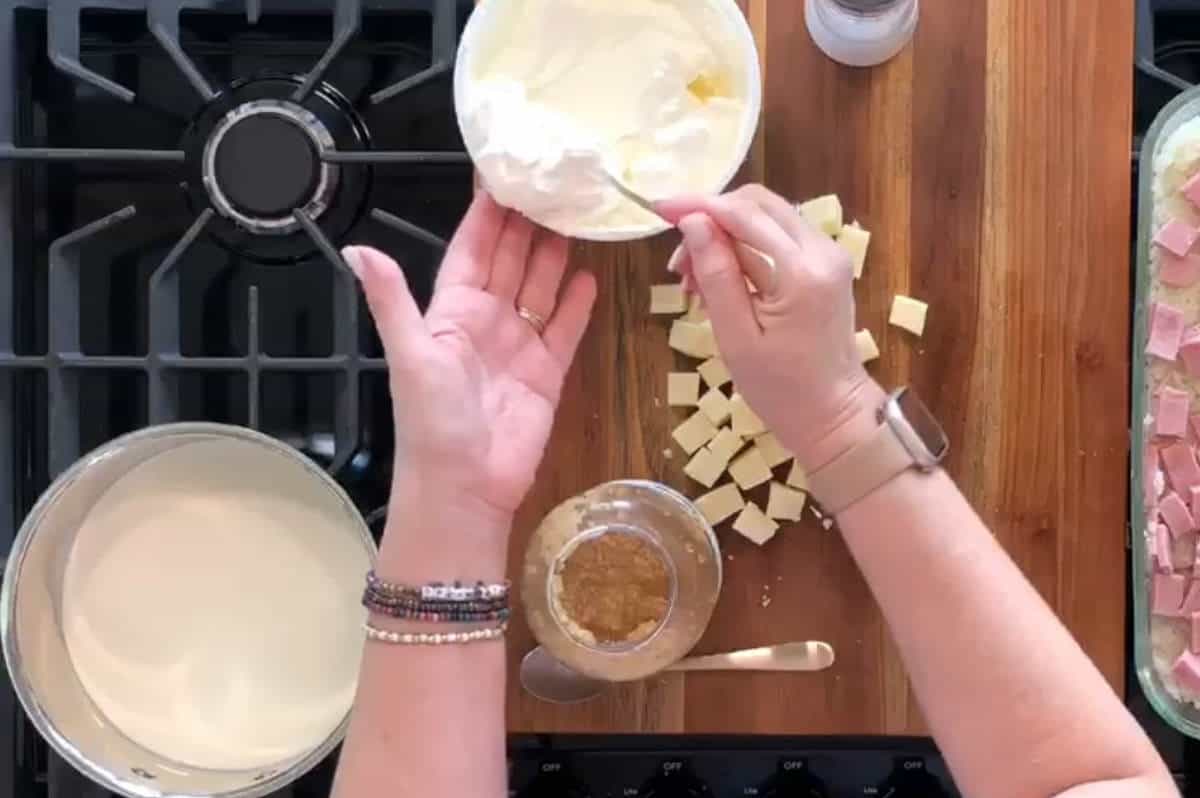 A person's hands preparing food in a kitchen. One hand holds a bowl with a spoon in it, containing a white creamy mixture, while the other hand scoops from the bowl. The scene includes a cutting board with cubed cheese and a jar of brown spice.