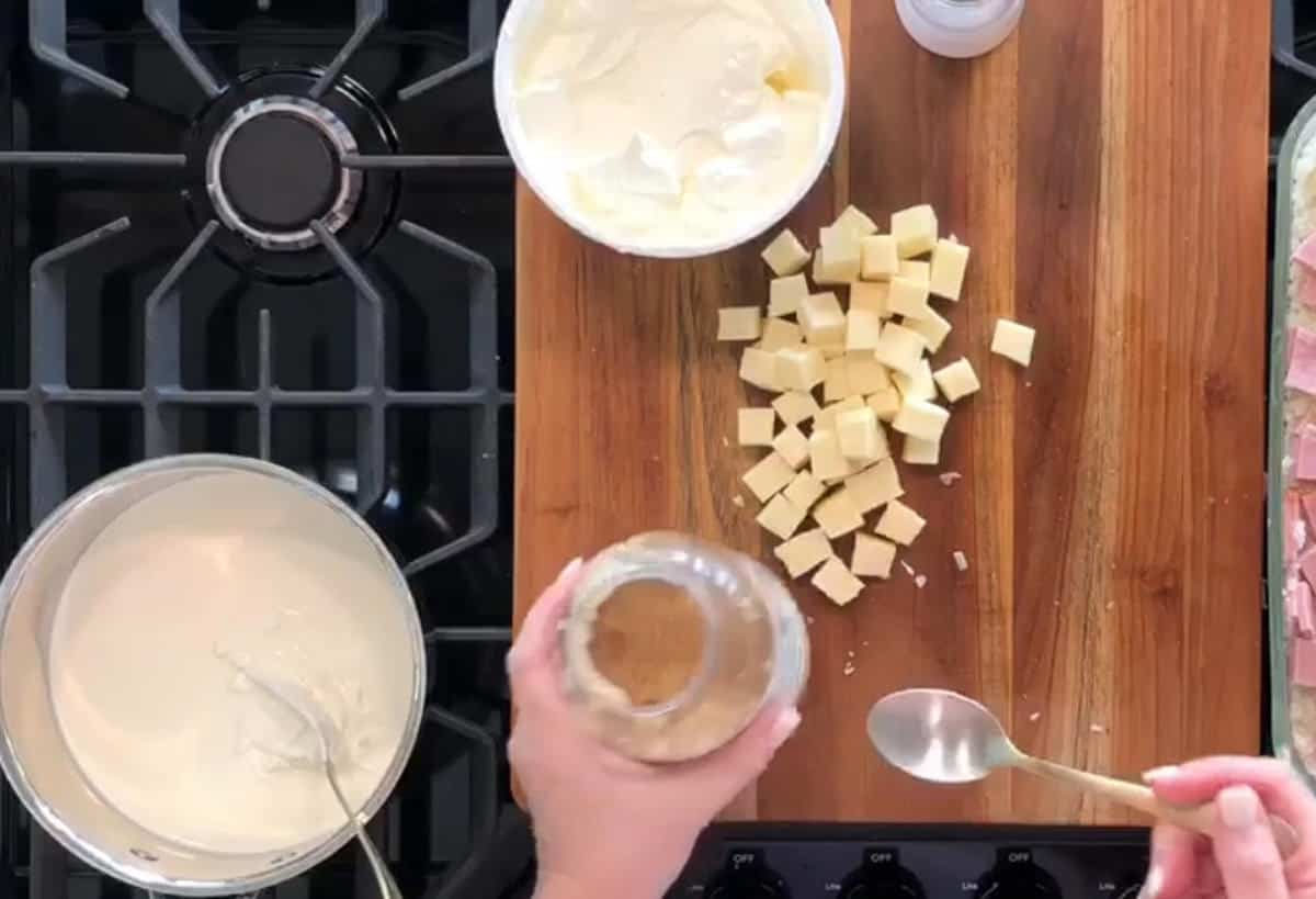 A stove with a pot of white sauce being stirred beside a cutting board. The cutting board has a bowl of white mixture, cubed cheese, and a person's hand holding a jar and a spoon. The person's other hand is holding a small spoon with some seasoning from the jar.