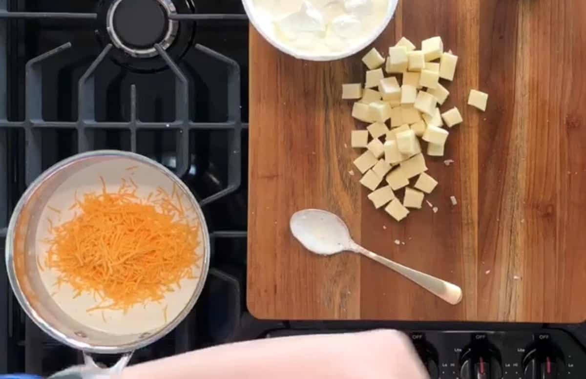 A wooden cutting board with cubed cheese and a bowl of sour cream sits next to a stovetop. A saucepan filled with shredded cheese is on the burner, and a spoon rests between the cutting board and the saucepan. A hand is seen at the bottom of the image.