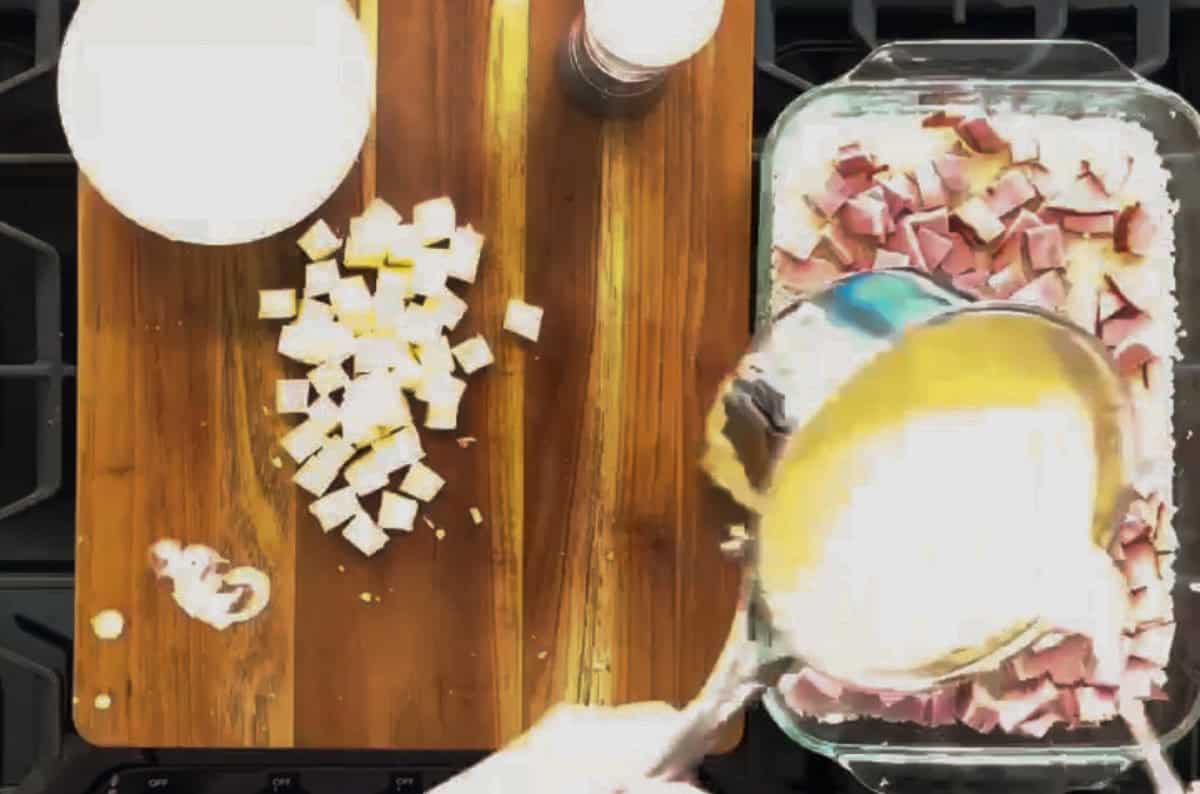 A person is pouring a creamy mixture over a dish filled with diced ham and other ingredients. The scene is from above, showcasing a wooden chopping board with cubed cheese, a bowl, and a salt shaker nearby.
