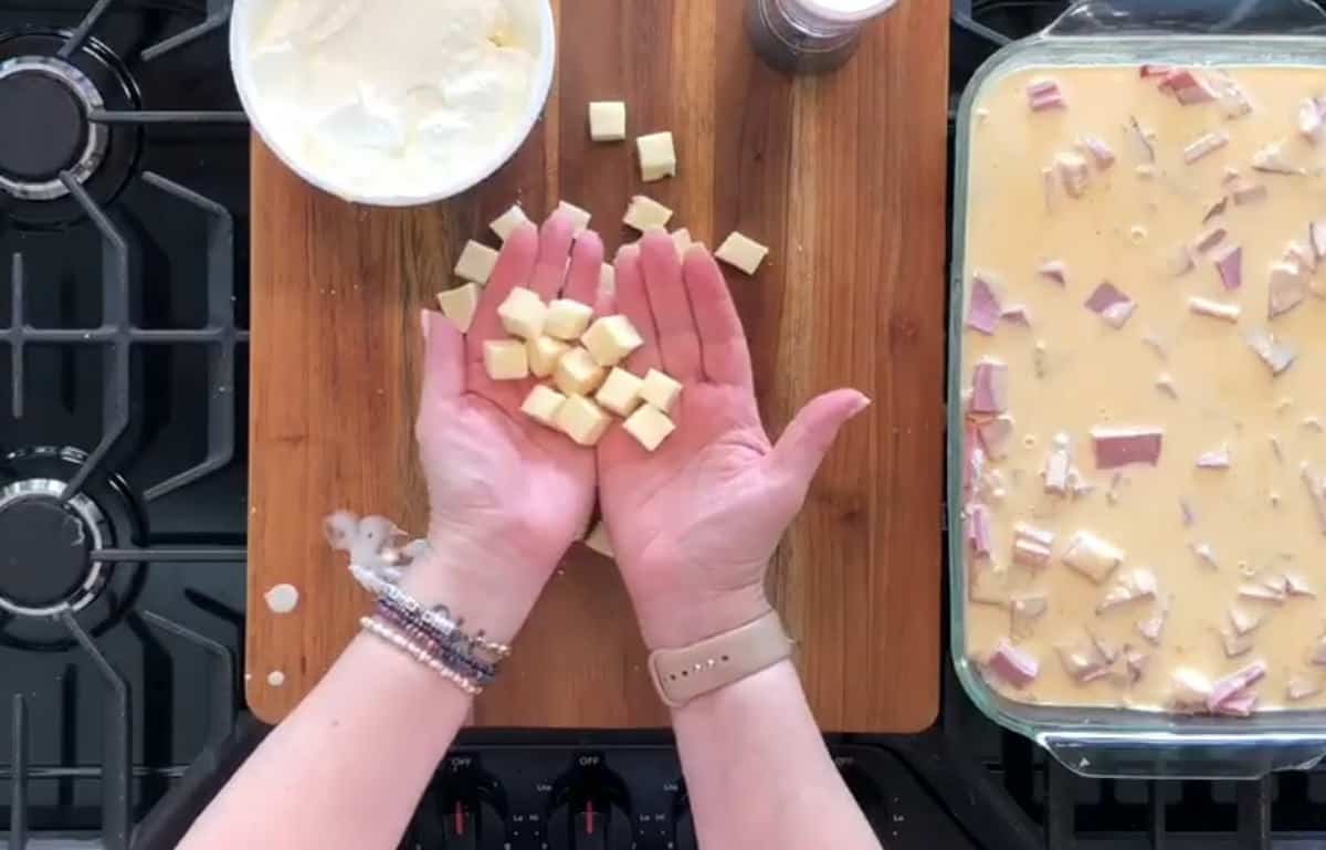 Two hands hold cubes of cheese over a wooden cutting board with a bowl of cream and a baking dish filled with a mixture of cream and bacon bits. The workspace is set on a stovetop.
