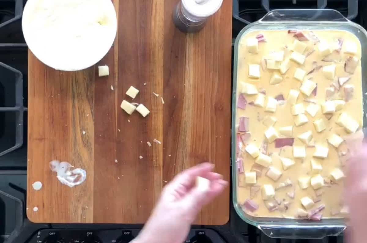 A wooden countertop with a cutting board is seen. On the cutting board, there is a bowl with cheese cubes and a small pile of cheese. To the right, there is a glass baking dish filled with a creamy mixture and cheese cubes. A hand is placing a cheese cube.